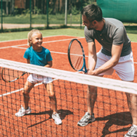 Cheerful father in sports clothing teaching his daughter to play tennis while both standing on tennis court