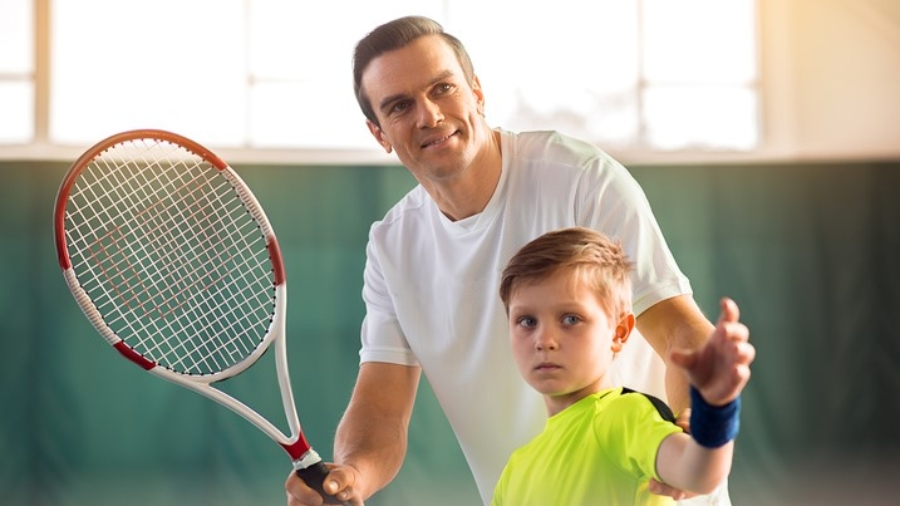Portrait of happy man teaching son how to hold racquet. He is putting his arms in right position and smiling. Boy is looking forward with concentration